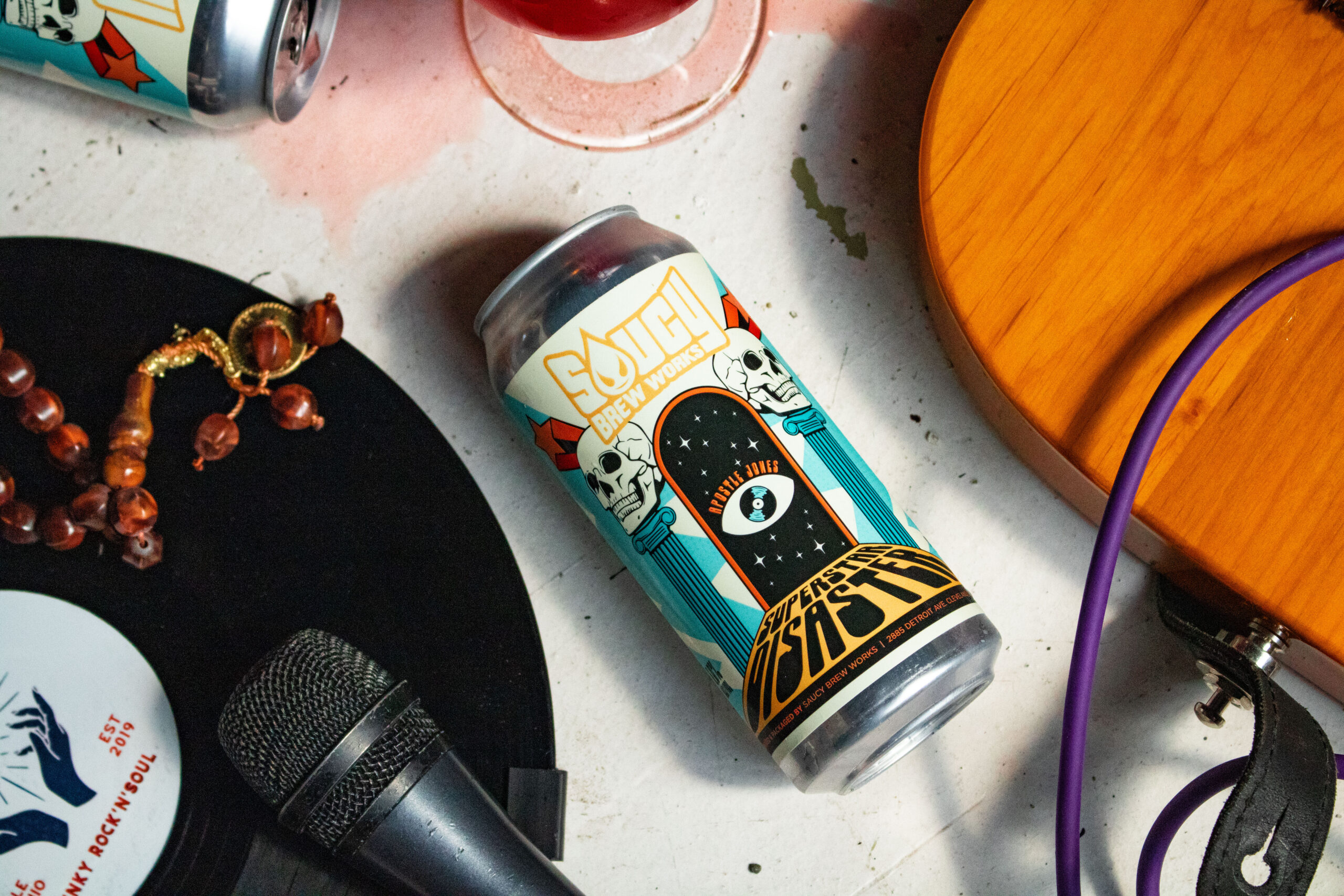 Beer can surrounded by spilled beer, a guitar, a microphone, and a record.